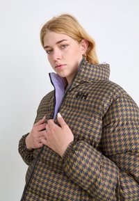 Houndstooth vzorovaná polstrovaná bunda v hnědé a béžové barvě, s vysokým límcem, zapínáním na zip a kontrastní levandulovou podšívkou.