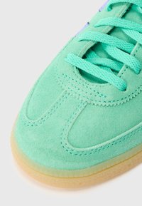 Gaiši zaļa zamšādas sporta kurpe ar saskaņotām auklām, noapaļotu purngalu un gumijas zoli. Ir izšūti detaļas un gluda tekstūra.