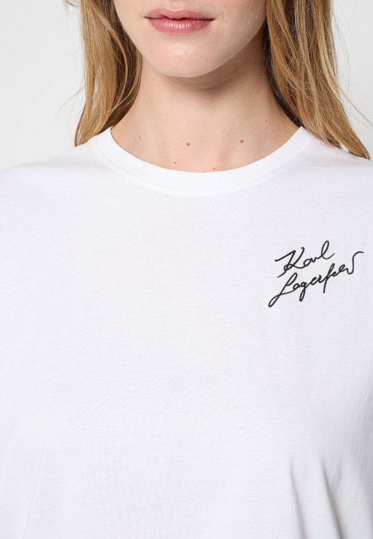 Weißes Baumwoll-T-Shirt mit Rundhalsausschnitt und kurzen Ärmeln, mit einem schwarzen, geschwungenen Logo auf der linken Brustseite. Glatte Textur.