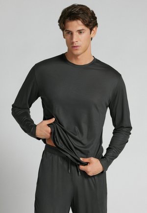 Mann in dunkelgrauem Langarmshirt und Hose hebt sein Shirt leicht an und blickt nach unten vor einfachem hellgrauem Hintergrund.