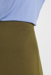 Ingrandimento di una persona che indossa un top azzurro chiaro infilato in una gonna o pantaloni verde oliva a vita alta.