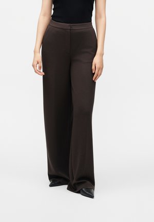 Femme portant un pantalon ample marron foncé à jambes larges avec des chaussures noires pointues et un top noir sans manches, debout devant un fond uni.