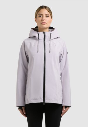 Femeie purtând o jachetă cu glugă gri deschis cu fermoar negru, stând în picioare pe un fundal simplu și luminos, privind înainte cu o expresie neutră.