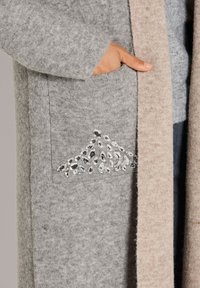 Hand in grauem Strickcardigan mit bestickter Tasche, die einen Cluster aus kleinen, klaren, facettierten Perlen zeigt, neben dem Rand eines beigen Strickbademantels oder -mantels.