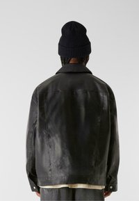 Schwarze Lederjacke mit glatter Textur, ausgestattet mit einem spitzen Kragen, Seitentaschen und einer entspannten Passform. Kombiniert mit einer navyfarbenen gerippten Mütze.