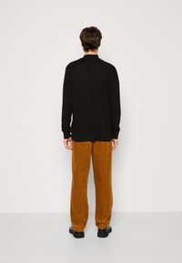 Black long-sleeve shirt with ribbed cuffs, paired with textured, loose-fitting, mustard-colored corduroy pants and black shoes.