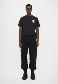 Zwarte katoen t-shirt met een neonkleurig geel logo op de linkerborst, gecombineerd met zwarte trainingsbroek met witte piping en zwarte laarzen.