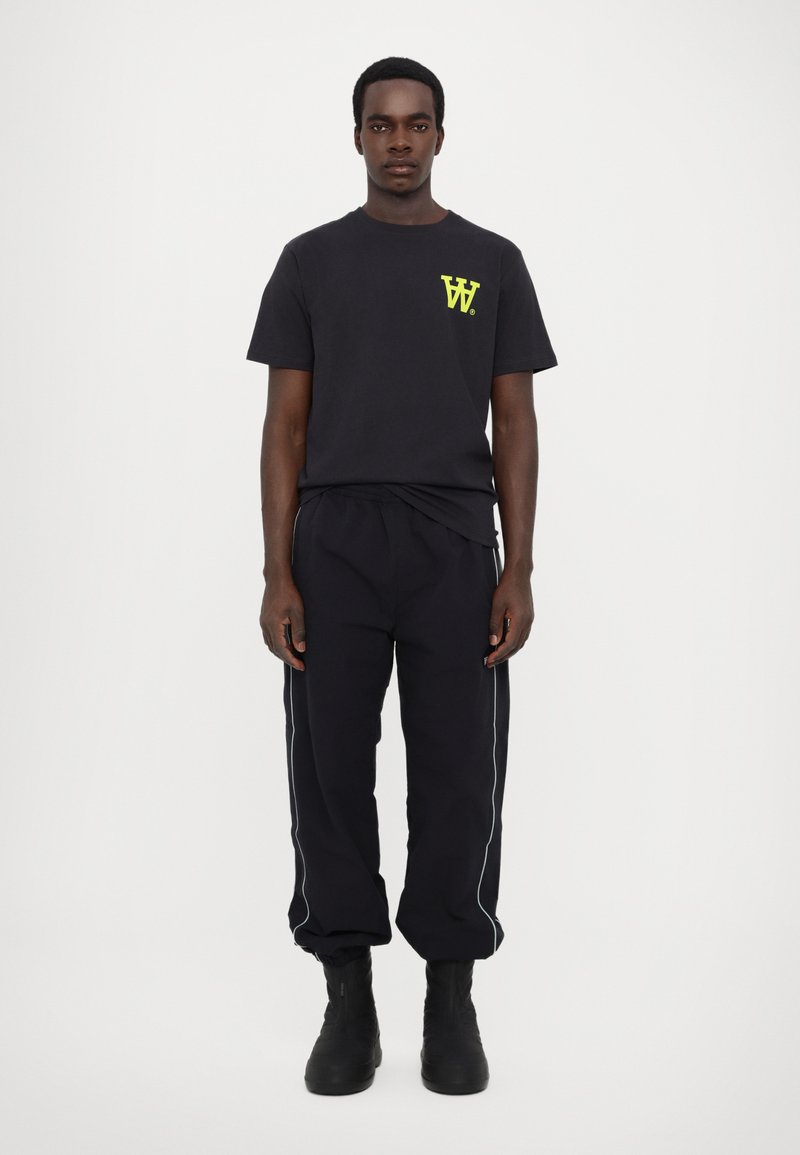 Zwarte katoen t-shirt met een neonkleurig geel logo op de linkerborst, gecombineerd met zwarte trainingsbroek met witte piping en zwarte laarzen.