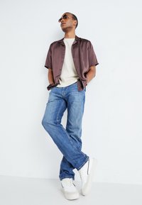 Chemise bordeaux à manches courtes, t-shirt beige, jeans bleus et baskets blanches avec des accents noirs. Le mannequin se tient devant un fond uni.