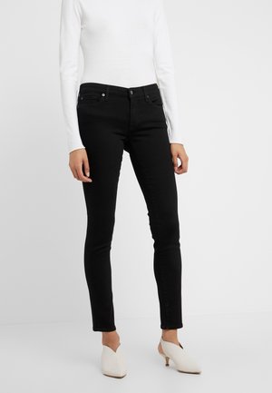 Kvinde iført tætsiddende sorte jeans, hvid langærmet top og hvide hælede slip-on sko, stående mod en ensfarvet hvid baggrund.