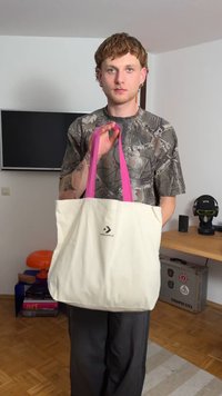 Platnena torba v krem barvi z rožnatimi ročaji, ki prikazuje Converse logo. Torba ima pravokotno obliko in je nestrukturirana, z mehko teksturo.