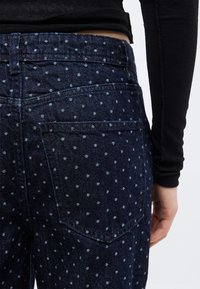 Άτομο που φοράει σκούρα μπλε τζιν με μικρές λευκές πουά και μαύρο μακρυμάνικο μπλουζάκι, δείχνοντας την πίσω τσέπη και το κάτω μέρος του κορμού.