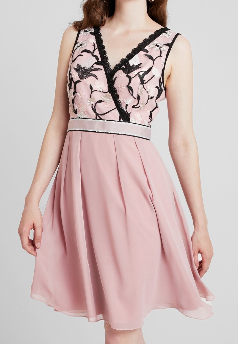 Bezramenné růžové šaty s černou květinovou výšivkou na korzetu, černým krajkovým lemem na výstřihu a bílým páskem v pase, které nosí stojící žena.