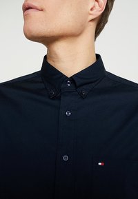 Camisa azul marino de botones con cuello, con botones negros, un bolsillo en el pecho con un pequeño detalle de marca y una textura suave de algodón.