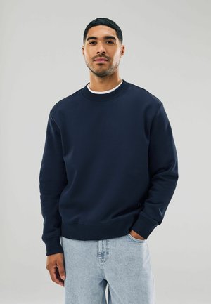 Ein Mann in einem marineblauen Sweatshirt und hellblauen Jeans steht vor einem neutralen Hintergrund und lächelt selbstbewusst.