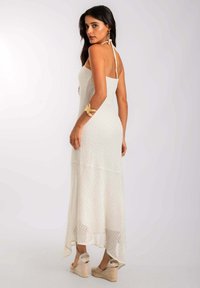 Vestido blanco de punto con tirantes halter, con un cuerpo ajustado y un dobladillo asimétrico, que presenta un sutil patrón texturizado. Combinado con sandalias de cuña beige.