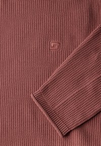 Ein pullover in rostfarbenem Strick mit einer strukturierten Rippenoptik, einem Rundhalsausschnitt und einem kleinen geprägt Logo auf der Brust.