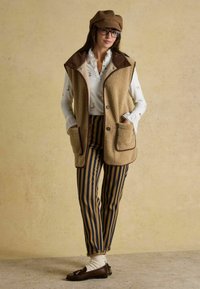 Beige Fleece-Weste mit braunen Akzenten und Taschen, kombiniert mit braunen und marineblauen gestreiften Hosen, braunen Slippern und einer passenden Mütze. Darunter ein weißes Hemd.