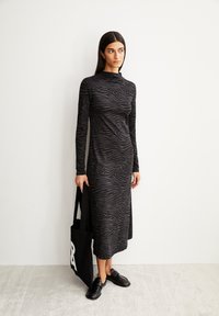 Robe noire à manches longues avec une texture semblable à celle d'un zèbre, fente sur le côté, associée à un sac fourre-tout noir et des chaussures noires à enfiler, sur une surface gris clair.