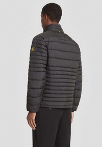 Zwarte pufferjack met horizontale gestikte textuur, hoge kraag en een patch op de linkermouw. Lichtgewicht met lange mouwen.