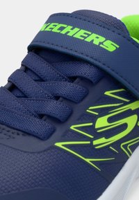 Tuvumā redzama tumši zilā Skechers sporta apava daļa ar laima zaļu logotipu, šņorēm un uzsūkšanas siksniņu, ar teksturētu audumu un baltu zoli.