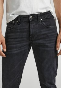 Černé džíny z denimu s přiléhavým střihem, pětice kapes, kovové zapínání a mírné odbarvení na přední straně. Hladká textura, žádné viditelné vzory.