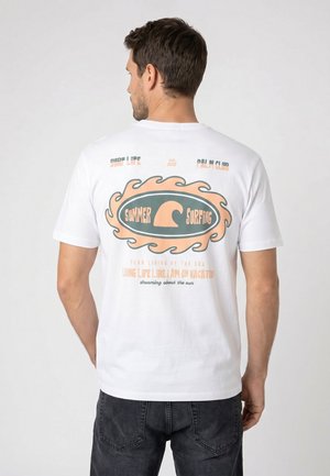 Man wearing white t-shirt with pastel orange and green surfing-themed graphic and text on back, paired with dark jeans.