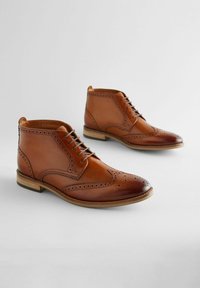 Bottines en cuir marron avec des détails brogues, un bout arrondi, des lacets et un talon en bois. Texture lisse avec une finition brillante.