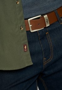 OLĪVU ZAĻA KREKLS AR PODIŅIEM, BRŪNA ĀDAS JOSTA AR SUDRABA SPĒCĪGU, UN TUMŠI ZILI DŽINSI AR ZELTA ŠUVĒM.