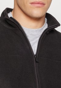 Svart fleece jakke med hel glidelås og høy krage, teksturert stoff, og kontrasterende grå indre lag synlig ved halslinningen.