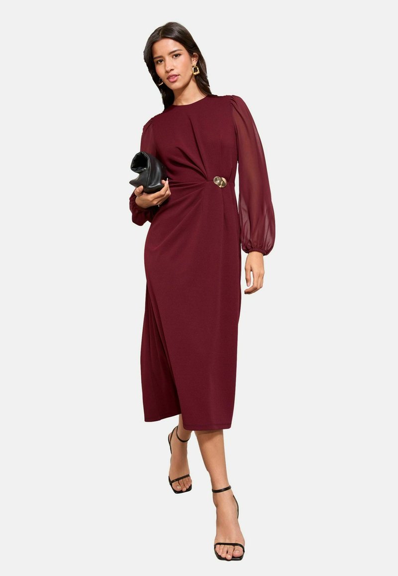 Mujer con un vestido midi burdeos de mangas largas transparentes, sosteniendo un bolso negro de mano y llevando sandalias negras de tiras, caminando hacia adelante.