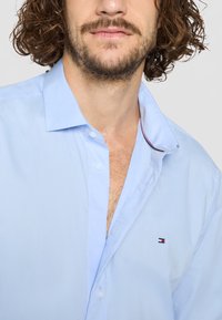 Lyseblå skjorte med knappestolpe og myk tekstur; har krage og en liten brodert logo i rødt, hvitt og blått nær brystet.