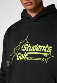Svart hettegenser med en stor, lys grønn grafikktekst som leser "Students Golf Performance Art" med stjerneakenter. Myk tekstur, avslappet design.
