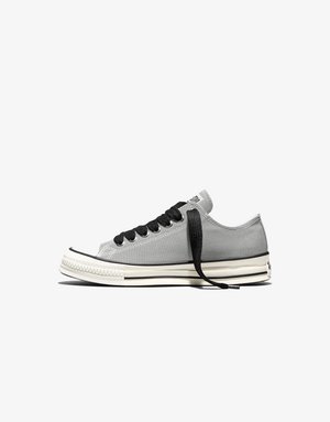 Laag gesneden grijze canvas sneaker met zwarte veters en een witte rubberen zool met zwarte strepen, zij-aanzicht op een witte achtergrond.