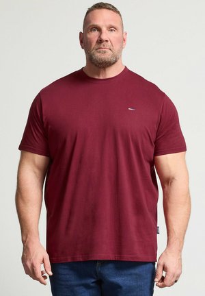 Gespierde man met kort haar en baard, gekleed in een effen bordeauxrood T-shirt met korte mouwen en blauwe spijkerbroek, staand tegen een lichte achtergrond.