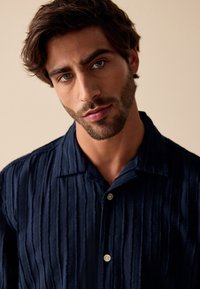 Marineblå tekstureret skjorte med lodrette striber, korte ærmer og en krave med knapper. Har to knapper foran.