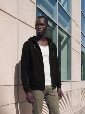 Homme portant un sweat à capuche noir et une chemise blanche, debout près d'un bâtiment moderne avec de grandes fenêtres, regardant sur le côté à la lumière du jour.