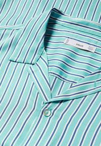 Chemise bleu clair à boutons avec rayures verticales blanches et bleu marine, posée à plat montrant le col et un seul bouton.