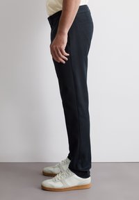 Pantaloni neri a gamba stretta realizzati in tessuto misto cotone, con tasche laterali, una texture liscia e indossati con scarpe da ginnastica bianche.