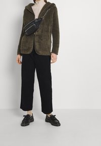 Olivengrøn, fuzzy hættejakke med forlommer, parret med sorte fløjl shorts og sorte sko med frynse-detaljer. Sort crossover taske.