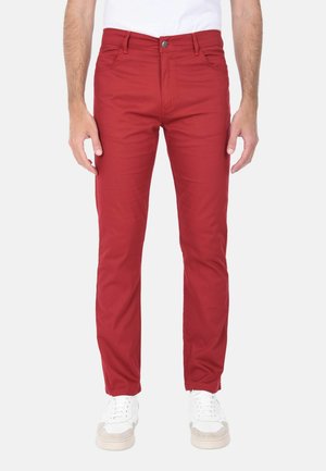 Uomo che indossa pantaloni rossi slim fit e sneakers bianche, in piedi davanti a uno sfondo bianco uniforme.
