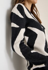 Musta-valkoinen kuvioitu neulepaita, jossa on neulottu pinta. Siinä on rohkeita geometrisia muotoja ja pyöreä pääntie sekä pitkät hihat.