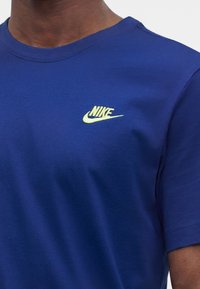 Άνδρας που φοράει βασιλικά μπλε μπλουζάκι Nike με μικρό κίτρινο λογότυπο Nike στο αριστερό στήθος σε απλό φόντο.