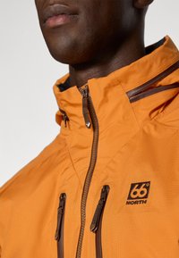 Oranje waterdichte jas heeft een hoge kraag, bruin accent bij de rits, een logo op de borst en dubbele ritszakken aan de voorkant.