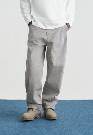 Personne portant un pantalon ample à rayures verticales, des bottes en daim beige et une chemise blanche à manches longues, les mains dans les poches, debout sur une moquette bleue.