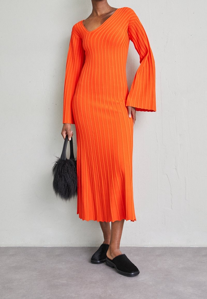 Robe midi orange côtelée avec manches évasées, col en V et finition texturée, associée à des chaussures noires sans lacets et un sac à main pelucheux.