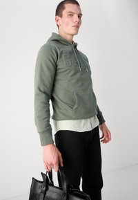 Sudadera verde oliva con bolsillo frontal y logo en relieve, superpuesta a una camisa clara, combinada con pantalones negros y un bolso negro.