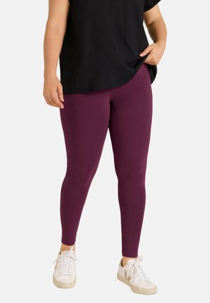 Persoon die paarse leggings, witte sneakers en een zwart top met korte mouwen draagt, met één hand in de zak, staand tegen een effen achtergrond.