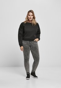 Felpa nera con dettagli a collo su spalle nere, abbinata a jeans skinny grigi. Il modello indossa sneakers alte nere. Sfondo semplice.
