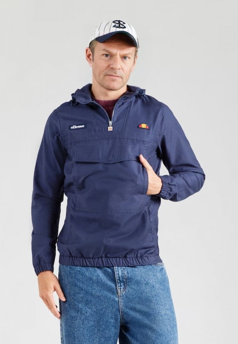 Giacca pullover blu navy con zip a tre quarti, dotata di tasca frontale e orlo elastico. Indossata con jeans blu chiaro e un cappellino bianco.